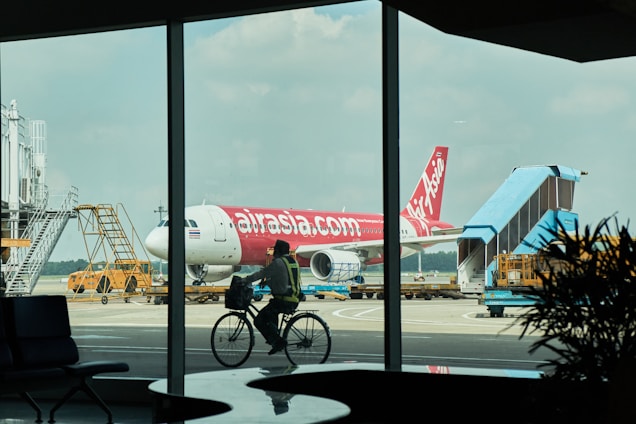 A red and white AirAsia airplane is parked at an airport gate. A worker in a safety vest is cycling in the foreground, silhouetted against the large glass windows of an airport terminal. The scene captures a busy airport environment with ground support equipment and a boarding stair vehicle nearby.