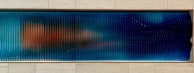 A horizontal panel with a series of vertical ridges that create a wavy texture. The surface displays a gradient of colors from deep blue on the right to a mix of blue and orange hues on the left. The panel is set within a light-colored, tiled wall.