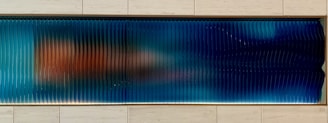 A horizontal panel with a series of vertical ridges that create a wavy texture. The surface displays a gradient of colors from deep blue on the right to a mix of blue and orange hues on the left. The panel is set within a light-colored, tiled wall.