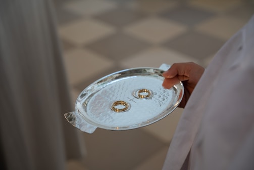 A close-up of a hand elegantly holding a textured silver tray that contains two gold rings. The tray appears to be held by someone wearing white garments, possibly a robe.