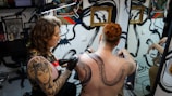 A tattoo artist is working on the back of a person with red hair, creating a large, intricate snake design. The studio has artistic wall decorations with black swirls and framed artwork. The artist has a detailed tattoo on their arm, and there is a mirror reflecting parts of the scene.