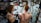 A tattoo artist is working on the back of a person with red hair, creating a large, intricate snake design. The studio has artistic wall decorations with black swirls and framed artwork. The artist has a detailed tattoo on their arm, and there is a mirror reflecting parts of the scene.