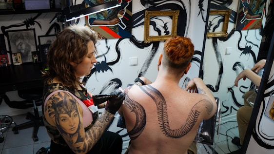 A tattoo artist is working on the back of a person with red hair, creating a large, intricate snake design. The studio has artistic wall decorations with black swirls and framed artwork. The artist has a detailed tattoo on their arm, and there is a mirror reflecting parts of the scene.
