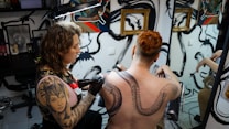 A tattoo artist is working on the back of a person with red hair, creating a large, intricate snake design. The studio has artistic wall decorations with black swirls and framed artwork. The artist has a detailed tattoo on their arm, and there is a mirror reflecting parts of the scene.