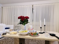 A cozy living room setting featuring a marble coffee table with various items including a bowl of grapes, a plate with watermelon slices, and a red rose arrangement in a clear vase. Two tall white candles are placed on black holders, and a glass of water and some snacks are present. The background shows sheer white curtains against a backdrop of a white sofa and a patterned carpet.