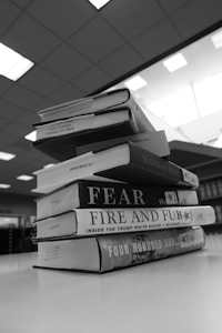 A stack of hardcover books is neatly arranged on a smooth surface in a library or bookstore setting. The titles are visible on the spines, with a fluorescent-lit ceiling above and bookshelves lining the background.