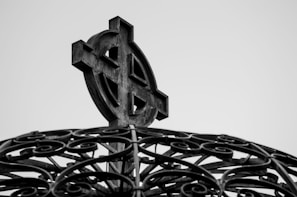A black and white close-up of an ornate Celtic cross mounted on a decorative base with intricate wrought iron swirls.