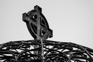 A black and white close-up of an ornate Celtic cross mounted on a decorative base with intricate wrought iron swirls.