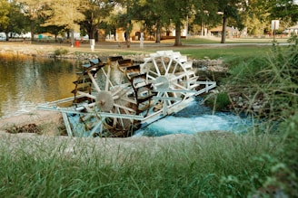 A white water wheel is situated in a serene park-like setting, partially submerged in a flowing stream. Surrounding the wheel are lush green grass and trees, with a calm pond reflecting the surroundings. Buildings and recreational areas are visible in the background, suggesting a peaceful, natural environment.