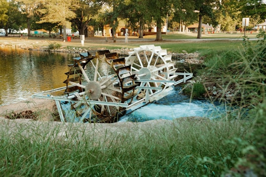 A white water wheel is situated in a serene park-like setting, partially submerged in a flowing stream. Surrounding the wheel are lush green grass and trees, with a calm pond reflecting the surroundings. Buildings and recreational areas are visible in the background, suggesting a peaceful, natural environment.
