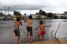 Three children are captured in mid-air as they jump into a body of water. They appear to be playing and having fun near a riverside with some buildings and houses visible in the background under a cloudy sky. The environment suggests a rural or village setting with lush greenery surrounding the area.