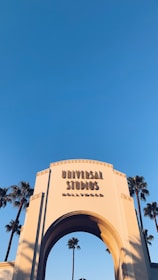 An iconic archway entrance to Universal Studios Hollywood with words prominently displayed against a clear blue sky. Tall palm trees are seen in the background, framing the structure.