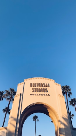An iconic archway entrance to Universal Studios Hollywood with words prominently displayed against a clear blue sky. Tall palm trees are seen in the background, framing the structure.