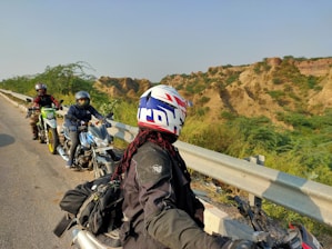 Three motorcyclists wearing helmets and protective gear are parked by the roadside. They are on a highway with a metal guardrail, and the backdrop features rugged hills covered in sparse vegetation. The sky is clear, indicating a sunny day.