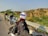 Three motorcyclists wearing helmets and protective gear are parked by the roadside. They are on a highway with a metal guardrail, and the backdrop features rugged hills covered in sparse vegetation. The sky is clear, indicating a sunny day.