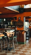A cozy pub setting featuring wooden chairs and tables on a tiled floor. The walls are adorned with Guinness signs, with warm lighting casting a comfortable ambiance. The large windows allow natural light to flood the space.