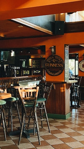 A cozy pub setting featuring wooden chairs and tables on a tiled floor. The walls are adorned with Guinness signs, with warm lighting casting a comfortable ambiance. The large windows allow natural light to flood the space.