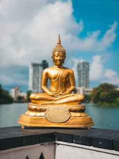 A golden Buddha statue sitting in a meditative pose on a pedestal, set against a background of a lake with modern buildings and greenery. The sky is partly cloudy and blue.