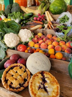 A colorful, abundant display of various fresh fruits and vegetables arranged on a wooden table. Visible items include leafy greens, cauliflower, tomatoes, peaches, radishes, carrots, broccoli, onions, cantaloupe, watermelon, and summer squash. Additionally, there are two homemade fruit pies with golden crusts placed prominently in the foreground.