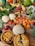 A colorful, abundant display of various fresh fruits and vegetables arranged on a wooden table. Visible items include leafy greens, cauliflower, tomatoes, peaches, radishes, carrots, broccoli, onions, cantaloupe, watermelon, and summer squash. Additionally, there are two homemade fruit pies with golden crusts placed prominently in the foreground.