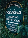 A sign surrounded by dense green foliage displays the text 'Agriturismo Révena Camping Guest House'. The background of the sign is dark green, with white lettering. It includes an arrow pointing to the left with additional welcoming messages and a phone number.