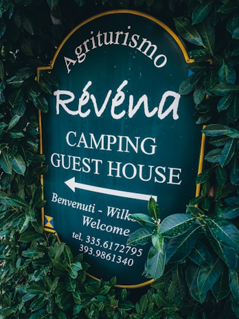 A sign surrounded by dense green foliage displays the text 'Agriturismo Révena Camping Guest House'. The background of the sign is dark green, with white lettering. It includes an arrow pointing to the left with additional welcoming messages and a phone number.