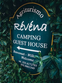 A sign surrounded by dense green foliage displays the text 'Agriturismo Révena Camping Guest House'. The background of the sign is dark green, with white lettering. It includes an arrow pointing to the left with additional welcoming messages and a phone number.