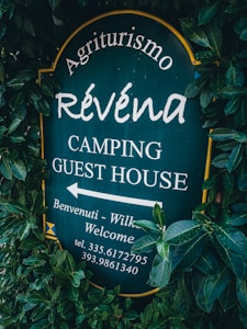 A sign surrounded by dense green foliage displays the text 'Agriturismo Révena Camping Guest House'. The background of the sign is dark green, with white lettering. It includes an arrow pointing to the left with additional welcoming messages and a phone number.