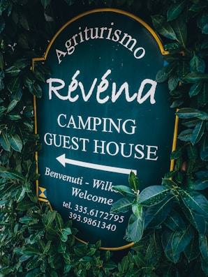 A sign surrounded by dense green foliage displays the text 'Agriturismo R&eacute;vena Camping Guest House'. The background of the sign is dark green, with white lettering. It includes an arrow pointing to the left with additional welcoming messages and a phone number.