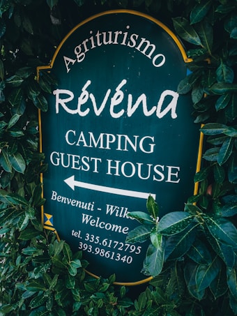 A sign surrounded by dense green foliage displays the text 'Agriturismo R&eacute;vena Camping Guest House'. The background of the sign is dark green, with white lettering. It includes an arrow pointing to the left with additional welcoming messages and a phone number.