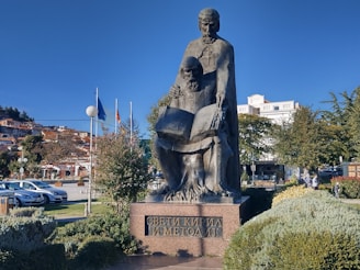 A stone statue depicting two historical figures, one standing and one seated, both holding an open book, placed in a landscaped park area. The background features a row of colorful flags, several buildings, and a clear blue sky.
