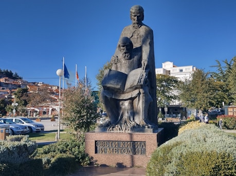 A stone statue depicting two historical figures, one standing and one seated, both holding an open book, placed in a landscaped park area. The background features a row of colorful flags, several buildings, and a clear blue sky.