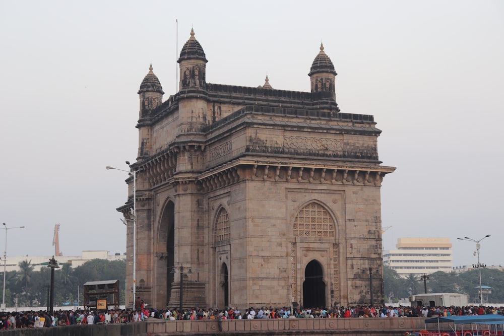 a large stone building with a crowd of people in front of it with Gateway of India in the background