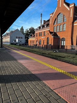 A train station platform alongside a red brick building with large arched windows and a chimney in the background. The platform is paved with red tiles and has a yellow tactile line running through the center. Overhead, there are electrical wires against a blue sky. In the distance, there are trees and additional buildings.
