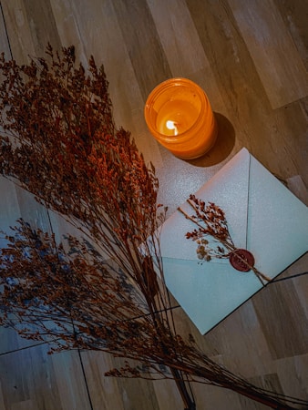 A warmly lit scene featuring a burning candle in a glass holder placed on a wooden surface. Next to the candle, there is a white envelope sealed with a red wax seal, embellished with small dried flowers. A cluster of dried plants adds an organic element to the composition.