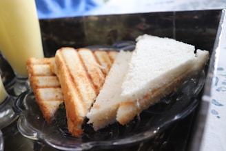 A glass plate holds a small selection of sandwiches, including a toasted sandwich with grill marks and a plain white bread sandwich cut into triangles. The sandwiches are placed on a reflective surface with a light-colored drink visible to the side.