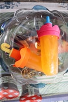 Two orange popsicles with colorful handles are placed in a transparent glass bowl. The popsicles have bright orange and pink tops, and the bowl sits on a table with patterned cloth. The background includes red and white checkered patterns and text that appears to be part of product labels.