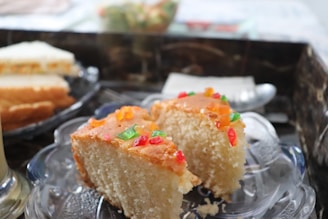 Two slices of a cake are placed on a glass plate. The cake appears to be a sponge cake with a light color and is topped with small colorful candied fruit pieces. The background includes additional blurred slices of cake and a hint of a table setting.