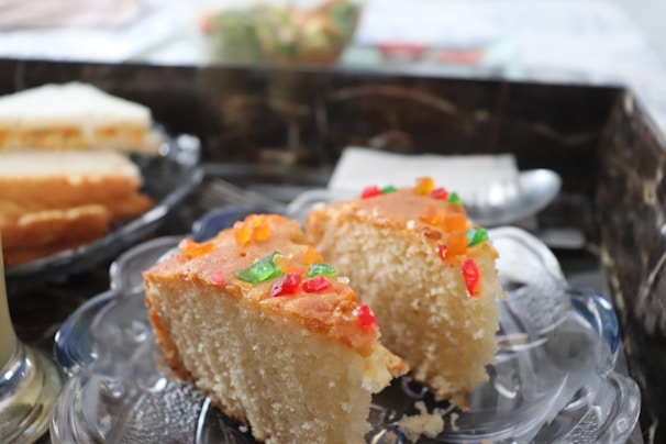 Two slices of a cake are placed on a glass plate. The cake appears to be a sponge cake with a light color and is topped with small colorful candied fruit pieces. The background includes additional blurred slices of cake and a hint of a table setting.