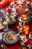 A richly decorated dining setup with a focus on culinary presentation. The main highlight is a dish featuring sushi rolls garnished with leafy greens on a dark plate. Nearby, a vibrant orange cocktail with slices of fruit sits next to a water glass with a lemon wedge. Kept company by a metallic lamp, a plate with bite-sized appetizers lies amidst decorative foliage.