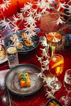 A richly decorated dining setup with a focus on culinary presentation. The main highlight is a dish featuring sushi rolls garnished with leafy greens on a dark plate. Nearby, a vibrant orange cocktail with slices of fruit sits next to a water glass with a lemon wedge. Kept company by a metallic lamp, a plate with bite-sized appetizers lies amidst decorative foliage.