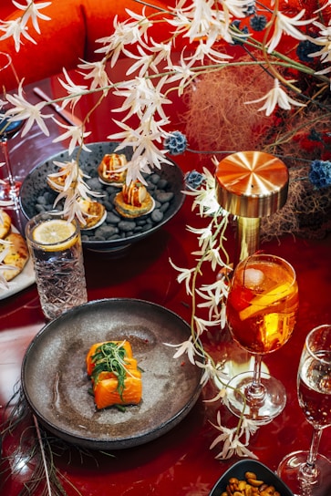 A richly decorated dining setup with a focus on culinary presentation. The main highlight is a dish featuring sushi rolls garnished with leafy greens on a dark plate. Nearby, a vibrant orange cocktail with slices of fruit sits next to a water glass with a lemon wedge. Kept company by a metallic lamp, a plate with bite-sized appetizers lies amidst decorative foliage.