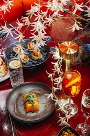 A richly decorated dining setup with a focus on culinary presentation. The main highlight is a dish featuring sushi rolls garnished with leafy greens on a dark plate. Nearby, a vibrant orange cocktail with slices of fruit sits next to a water glass with a lemon wedge. Kept company by a metallic lamp, a plate with bite-sized appetizers lies amidst decorative foliage.