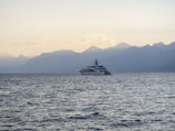 A large yacht is sailing on calm ocean waters with a backdrop of hazy, mountainous terrain. The sky is clear with soft, ambient lighting suggesting either dawn or dusk.