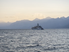 A large yacht is sailing on calm ocean waters with a backdrop of hazy, mountainous terrain. The sky is clear with soft, ambient lighting suggesting either dawn or dusk.
