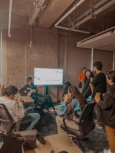 A group of people in an office setting appears to be engaged in a team meeting or presentation. Several individuals are seated around a large screen, and one person seems to be explaining something on the screen. The atmosphere is collaborative with participants using laptops and notepads.