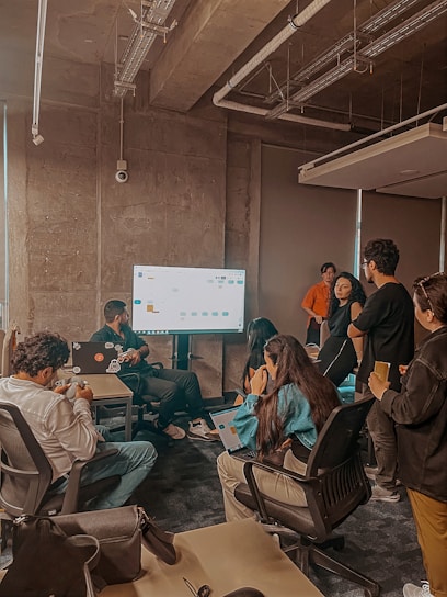 A group of people in an office setting appears to be engaged in a team meeting or presentation. Several individuals are seated around a large screen, and one person seems to be explaining something on the screen. The atmosphere is collaborative with participants using laptops and notepads.