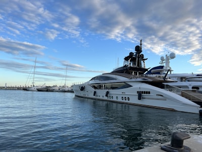 A luxurious yacht is docked at a marina under a partly cloudy sky. The yacht features sleek design elements with white hull and dark accents. Several other boats are also moored nearby, and there is a visible pier in the background.