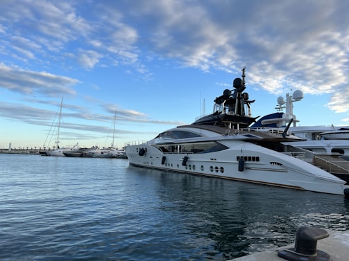 A luxurious yacht is docked at a marina under a partly cloudy sky. The yacht features sleek design elements with white hull and dark accents. Several other boats are also moored nearby, and there is a visible pier in the background.