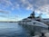 A luxurious yacht is docked at a marina under a partly cloudy sky. The yacht features sleek design elements with white hull and dark accents. Several other boats are also moored nearby, and there is a visible pier in the background.